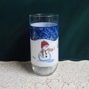 May include: A clear glass with a winter-themed design. The glass features a blue band with a snowy pattern at the top and a snowman graphic in the center. The snowman wears a red hat and scarf.