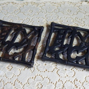 May include: Two black cast iron hot pads with a decorative design. The hot pads are square shaped and have a raised design that looks like bamboo.