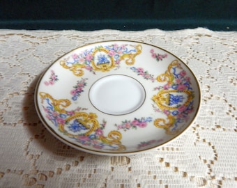May include: A white porcelain saucer with a gold rim and a floral pattern in shades of pink, blue, and yellow. The pattern features ornate scrolls and a central motif of a vase with flowers.