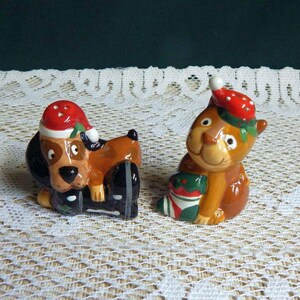 May include: A pair of ceramic salt and pepper shakers shaped like a brown dog and a brown cat. Both animals wear red Santa hats with white dots. The dog is lying down, and the cat is sitting upright while holding a green and red Christmas stocking.