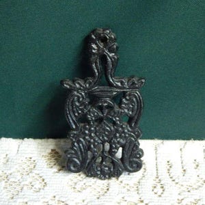 May include: A black cast iron trivet featuring an intricate openwork design with scrolling leaf patterns and a central cluster of grapes. The rectangular piece includes a small circular hole at the top for hanging and a textured, matte finish.