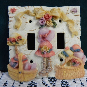 May include: A white ceramic light switch cover with a 3D design featuring two ladies wearing hats and baskets of flowers. The design is in shades of pink, yellow, and green.