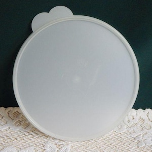 May include: A round, white plastic lid with a small, heart-shaped handle. The lid is slightly translucent and appears to be for a container. The background is a dark green color.