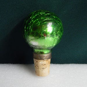 May include: Green glass bottle stopper with a cork base. The glass is textured and has a metallic sheen.