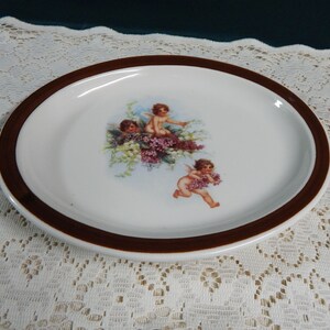 May include: A white ceramic plate with a brown rim features a vintage illustration of three cherubs with flowers. The cherubs are depicted with wings and are holding flowers.