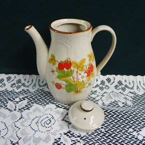 May include: A ceramic teapot with a floral design featuring strawberries and leaves. The teapot is beige with brown accents and a lid.