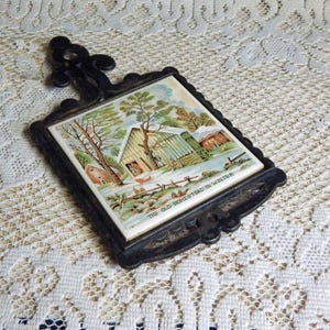 May include: A black cast iron trivet featuring a square ceramic tile insert with a winter landscape illustration of a house and trees. The text THE OLD HOMESTEAD IN WINTER is printed at the bottom of the tile. The metal frame has a decorative handle.