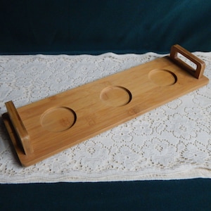 May include: A long, rectangular wooden serving tray with three circular indentations. The tray has a handle on each end and is made of light brown bamboo.