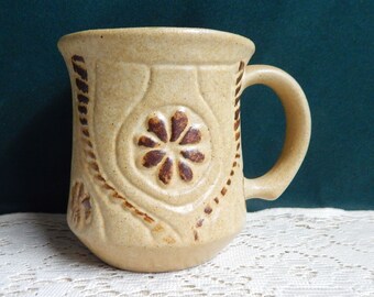 May include: Tan ceramic mug with a handle. The mug features a raised floral design in the center and a decorative border. The handle is curved and the mug has a slightly flared rim. The mug is a neutral color.