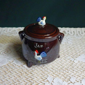 May include: A brown ceramic jam jar with a lid, featuring a rooster figurine on top. The jar has handles and a rooster illustration with the word "Jam" written on the front. The jar is on a white lace tablecloth.