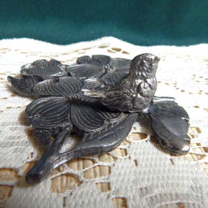 May include: A silver metal figurine of a bird perched on a branch with three stylized dogwood blossoms. The figurine is detailed with textured leaves and flowers.