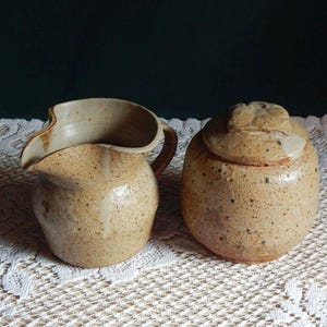 May include: A handmade ceramic creamer and lidded sugar bowl set. The pottery is a speckled tan color with a rustic, organic shape. The creamer has a curved spout and handle. The sugar bowl has a lid with a small knob. The set is displayed on a white lace tablecloth.