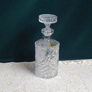 May include: A clear glass decanter with a faceted design and a stopper. The decanter has a label that reads "Lead Crystal Made in Czechoslovakia".