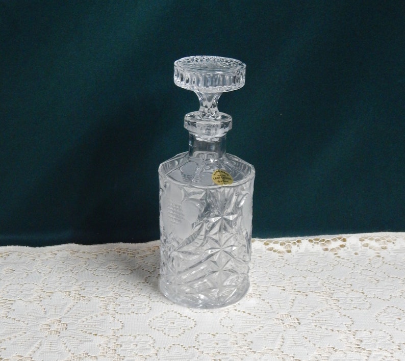 May include: A clear glass decanter with a faceted design and a stopper. The decanter has a label that reads "Lead Crystal Made in Czechoslovakia".