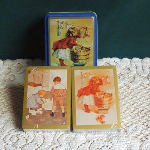 May include: A vintage playing card set featuring illustrations of children at play. The set includes two decks of cards and a blue tin with a matching image. The cards and tin feature a charming, nostalgic aesthetic.