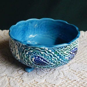 May include: A turquoise ceramic bowl with a scalloped rim and three small feet. The bowl features a detailed paisley pattern in shades of blue, green, and white. The interior is a solid, glossy turquoise.