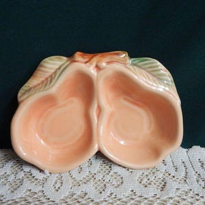 May include: A ceramic pear-shaped dish with two compartments. The dish is peach-colored with green and orange accents on the leaves. The dish is likely used for serving snacks or as a decorative piece.