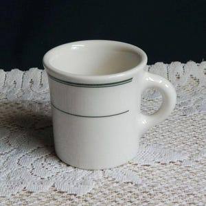 May include: A white ceramic mug with a rounded handle. The mug has a dark green stripe near the rim and another below the rim. The mug is sitting on a white lace tablecloth.