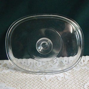 May include: A clear, oval-shaped glass lid featuring a central knob handle and a radiating sunburst pattern of fine, etched lines extending from the center toward the smooth, raised outer rim of the lid, designed for a matching glass dish.