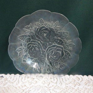 May include: Clear glass serving plate with a scalloped edge. The plate features an embossed design of three roses surrounded by leaves. The floral pattern is centered on the plate. The plate is on a white lace surface.