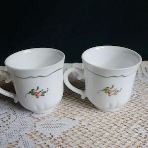 May include: Two white ceramic mugs with a floral design. Each mug has a curved handle and a scalloped green line near the rim. A small floral motif with red and green accents is painted on the front of each mug.
