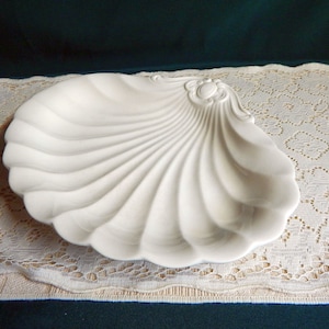 May include: A white ceramic serving dish shaped like a seashell. The dish has a smooth, glossy finish and a slightly raised rim.