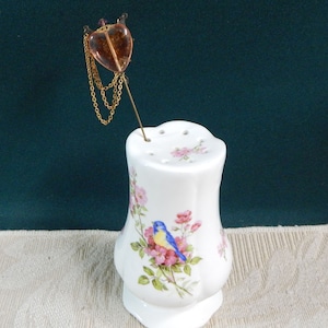 May include: White ceramic pincushion with a floral design and a blue bird. A gold heart-shaped pin with a chain is stuck in the pincushion.