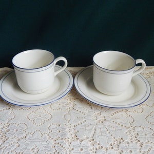 May include: Two white ceramic teacups with blue trim, each on a matching saucer. The cups and saucers have a simple, elegant design.