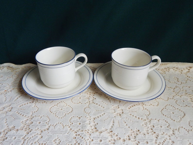 May include: Two white ceramic teacups with blue trim, each on a matching saucer. The cups and saucers have a simple, elegant design.