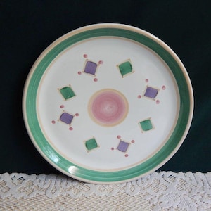 May include: White ceramic plate with a green and tan border. The plate features a central pink and yellow spiral design, surrounded by green and purple diamond shapes and small pink dots. The plate is on a white lace tablecloth.