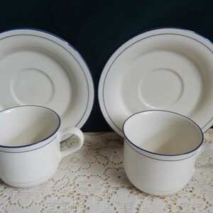 May include: Two white ceramic teacups with blue trim and matching saucers. The cups and saucers have a simple design with a single blue line around the rim.