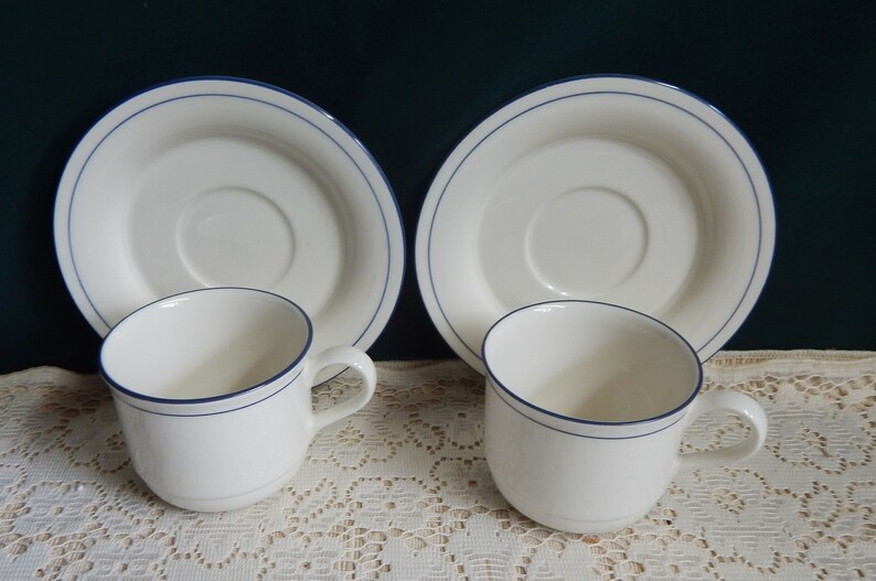 May include: Two white ceramic teacups with blue trim and matching saucers. The cups and saucers have a simple design with a single blue line around the rim.