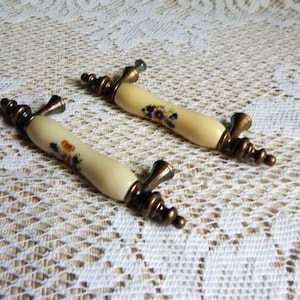May include: Two vintage cabinet pulls with a cream-colored ceramic handle and bronze-toned metal hardware. The handles are decorated with a floral pattern in shades of blue, orange, and purple. These pulls are ideal for adding a touch of vintage charm to furniture.