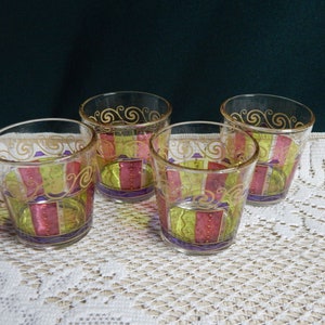 May include: Four clear glass tumblers with a decorative design. The glasses have a yellow, pink, and purple striped pattern with gold accents.