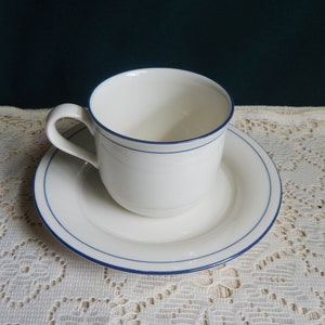 May include: A white ceramic teacup and saucer with a thin blue trim. The cup has a handle and the saucer has a slightly raised edge.