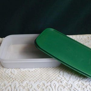 May include: A rectangular food storage container made of translucent white plastic with a matching dark green plastic lid. The container has rounded corners and a flat base, while the lid is positioned at an angle partially covering the top.