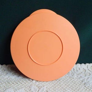 May include: A round, peach-colored plastic lid with a raised handle and a circular indentation in the center. The lid is designed to fit a container and is likely used for food storage. The lid is made of durable plastic.