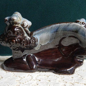 May include: A brown glazed ceramic figurine of a mythical creature with a curled tail and a head with a crown. The figurine has a blue and white glaze on the back and is sitting on a white lace surface.