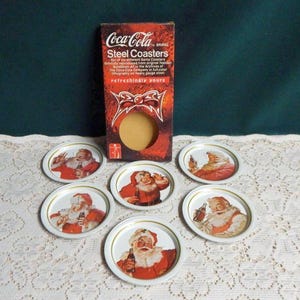 May include: Set of six Coca-Cola brand steel coasters featuring Santa Claus images. Each coaster has a different image of Santa Claus with a Coca-Cola bottle. The coasters are in a box with the Coca-Cola logo and the text "refreshingly yours".