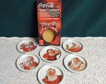 May include: Set of six Coca-Cola brand steel coasters featuring Santa Claus images. Each coaster has a different image of Santa Claus with a Coca-Cola bottle. The coasters are in a box with the Coca-Cola logo and the text "refreshingly yours".