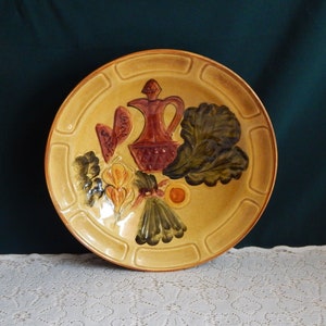 May include: A ceramic plate with a raised design of a pitcher, vegetables, and fruit. The plate is decorated with brown, green, yellow, and red paint.