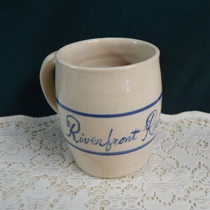 May include: A white ceramic mug with a blue band and the words "Riverfront Restaurant" in blue script.