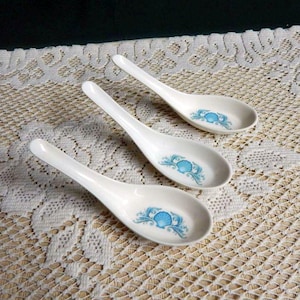 May include: Three white ceramic soup spoons with a blue floral design. The spoons are arranged on a white lace tablecloth. The spoons have a long handle and a rounded bowl, perfect for serving soup or other dishes.