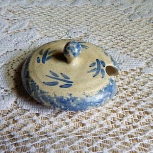 May include: A blue and white ceramic lid with a small spout and a knob on top. The lid has a simple leaf design.