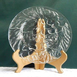 May include: A clear glass plate with a grape and leaf design, displayed on a gold-colored stand. The plate's embossed pattern features clusters of grapes and detailed leaves. The stand has a three-legged design.
