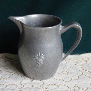 May include: A silver pitcher with a pineapple design. The pitcher is made of metal and has a smooth, shiny finish.
