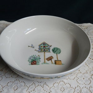 May include: A white ceramic bowl with a birdhouse and trees design. The birdhouse is blue and green with red hearts. The trees are green and have brown trunks. The bowl has a yellow and green border.