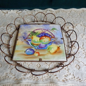 May include: A vintage ceramic tile with a hand-painted image of a bowl of fruit, including grapes, pears, and apples. The tile is set in a decorative metal frame.