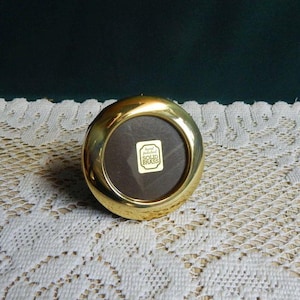 May include: A circular, polished gold-tone brass frame with a smooth, rounded edge. The center features a dark brown circular insert with a small, rectangular white sticker that reads "hand polished SOLID BRASS" in black text.