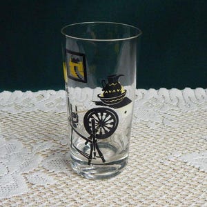 May include: A clear glass with black and yellow illustrations of a spinning wheel, a pitcher, and a framed picture. The glass is cylindrical and sits on a white lace tablecloth. The background is a dark green color.